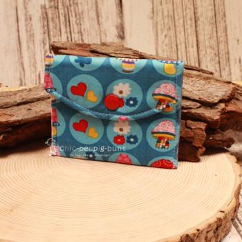 Kleiner handgenähter Geldbeutel von chic-peppig-bunt aus blauem Stoff mit Herzen und Blumen sowie rot-weiß kariertem Futter auf hellem Hintergrund.