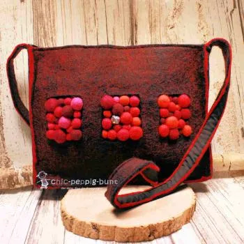 Handgefertigte graue Filztasche mit aufgenähten Bommeln in Pink, Rot und Lila sowie einer silbernen Metallperle vor hellem Hintergrund.
