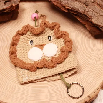 Gehäkelter Löwe Schlüsselanhänger in Beige mit brauner Mähne, grüner Schnur und rosa Perle auf einer Holzscheibe.