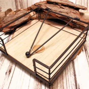 Quadratischer Serviettenhalter aus schwarzem Metall-Zaun und hellem Holz mit einem Kugel-Gewichtarm auf einer Baumscheibe.