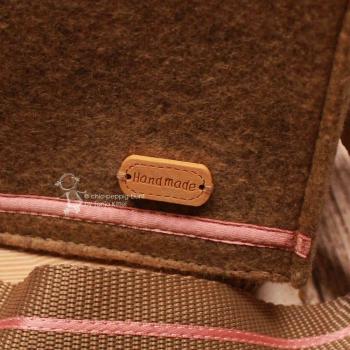 Nahaufnahme des aufgenähten, ovalen Leder-Labels mit der feinen Prägung Hand made auf dem beigen Filzstoff.