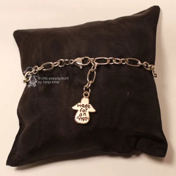 Versilbertes Bettelarmband aus Metall mit Schutzengel-Anhänger Schriftzug made for an Angel auf braunem Schmuckkissen.