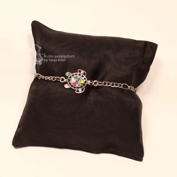 : Versilbertes Gliederarmband mit bunter Schildkröte präsentiert auf einem schwarzen Schmuckkissen.