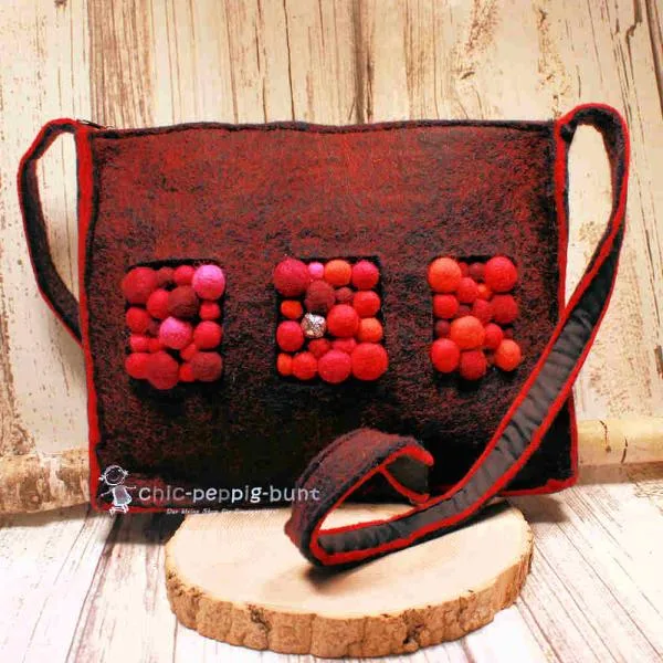 Handgefertigte graue Filztasche mit aufgenähten Bommeln in Pink, Rot und Lila sowie einer silbernen Metallperle vor hellem Hintergrund.