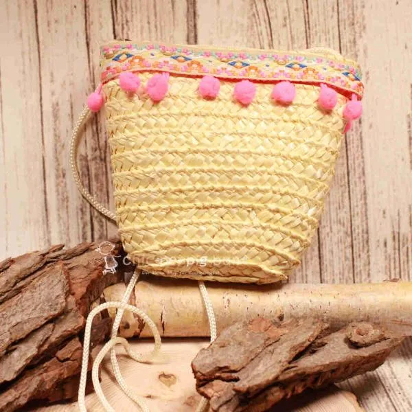 Kleine runde Korbtasche für Kinder aus naturfarbenem Rattan mit rosa Bommeln, heller Bordüre und Stoff-Innenfutter.
