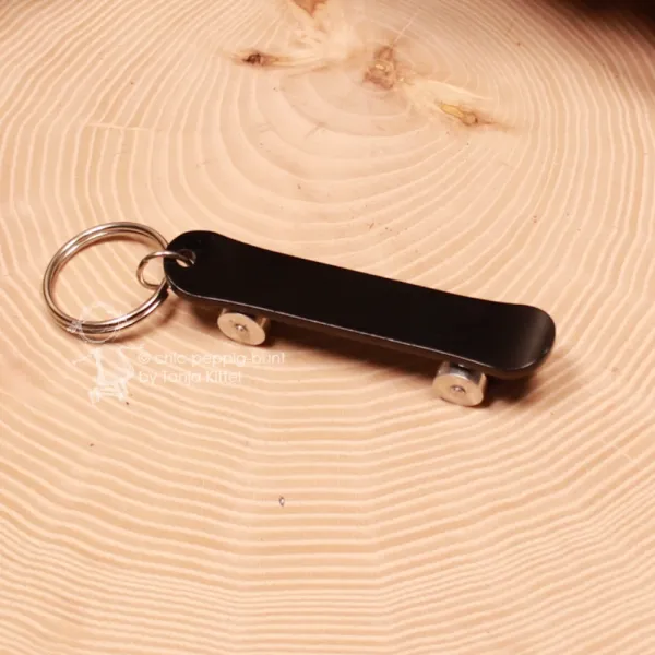 Schwarzer Skateboard Schlüsselanhänger aus Metall mit silbernen Reifen auf einer Holzscheibe liegend.
