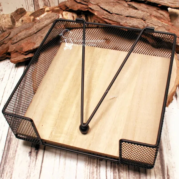 Quadratischer Serviettenhalter aus schwarzem Metall-Gitter mit hellem Holzboden und Kugel-Beschwerer auf einer Baumscheibe.