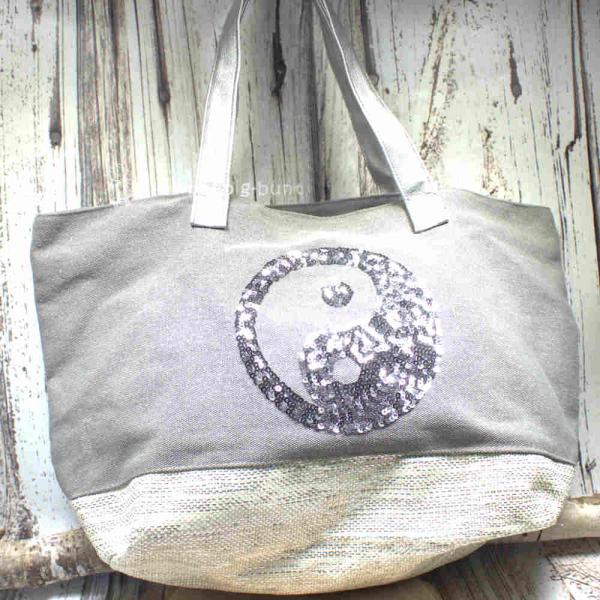 Graue Shoppertasche mit silbernem Yin Yang Pailletten-Motiv, weißem Boden mit Silberstreifen und grauen Kunstleder-Griffen vor Holz-Hintergrund.