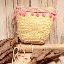 Kleine runde Korbtasche für Kinder aus naturfarbenem Rattan mit rosa Bommeln, heller Bordüre und Stoff-Innenfutter.