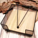 Quadratischer Serviettenhalter aus schwarzem Metall-Gitter mit hellem Holzboden und Kugel-Beschwerer auf einer Baumscheibe.